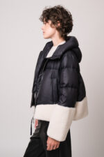 Model trägt eine schwarze Daunenjacke mit weißem Fellbesatz der Marke Duno, einen beigen Pullover der Marke Herzens und eine schwarze Hose der Marke Cambio.