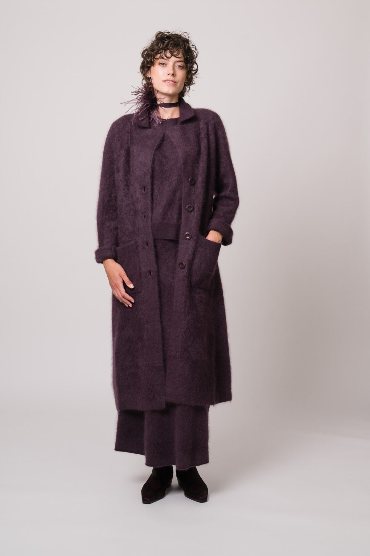 Model trägt einen Strickmantel, einen Pullover und einen Rock in violett der Marke FFC sowie einen Federschmuck der Marke Susanne Bommer.