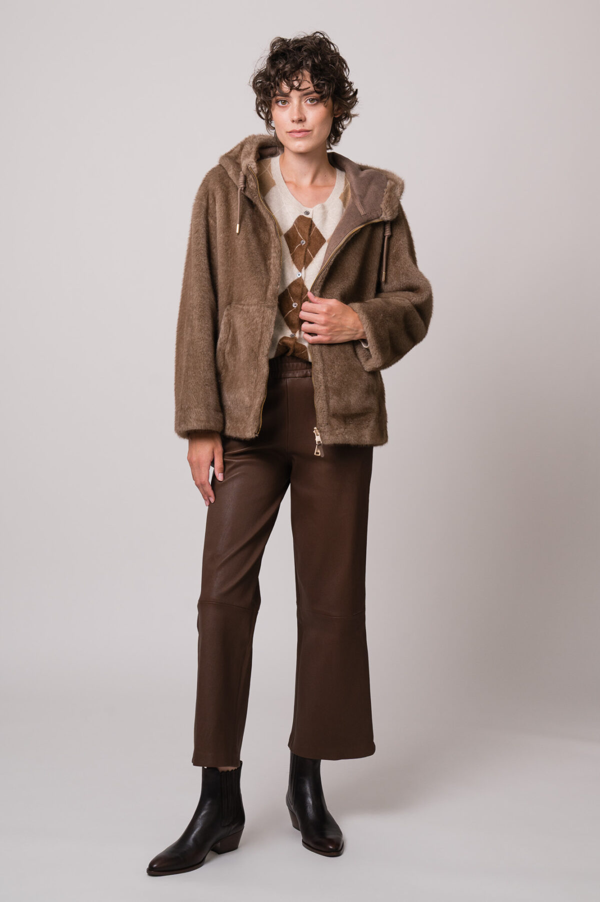 Model trägt eine beide Fake Fur Jacke der Marke Mabrun, eine beige Strickweste mit Rautenmuster der Marke The Mercer und eine braune Lederhose der Marke Hemisphere.