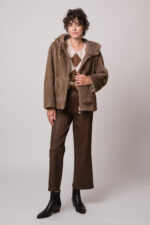 Model trägt eine beide Fake Fur Jacke der Marke Mabrun, eine beige Strickweste mit Rautenmuster der Marke The Mercer und eine braune Lederhose der Marke Hemisphere.