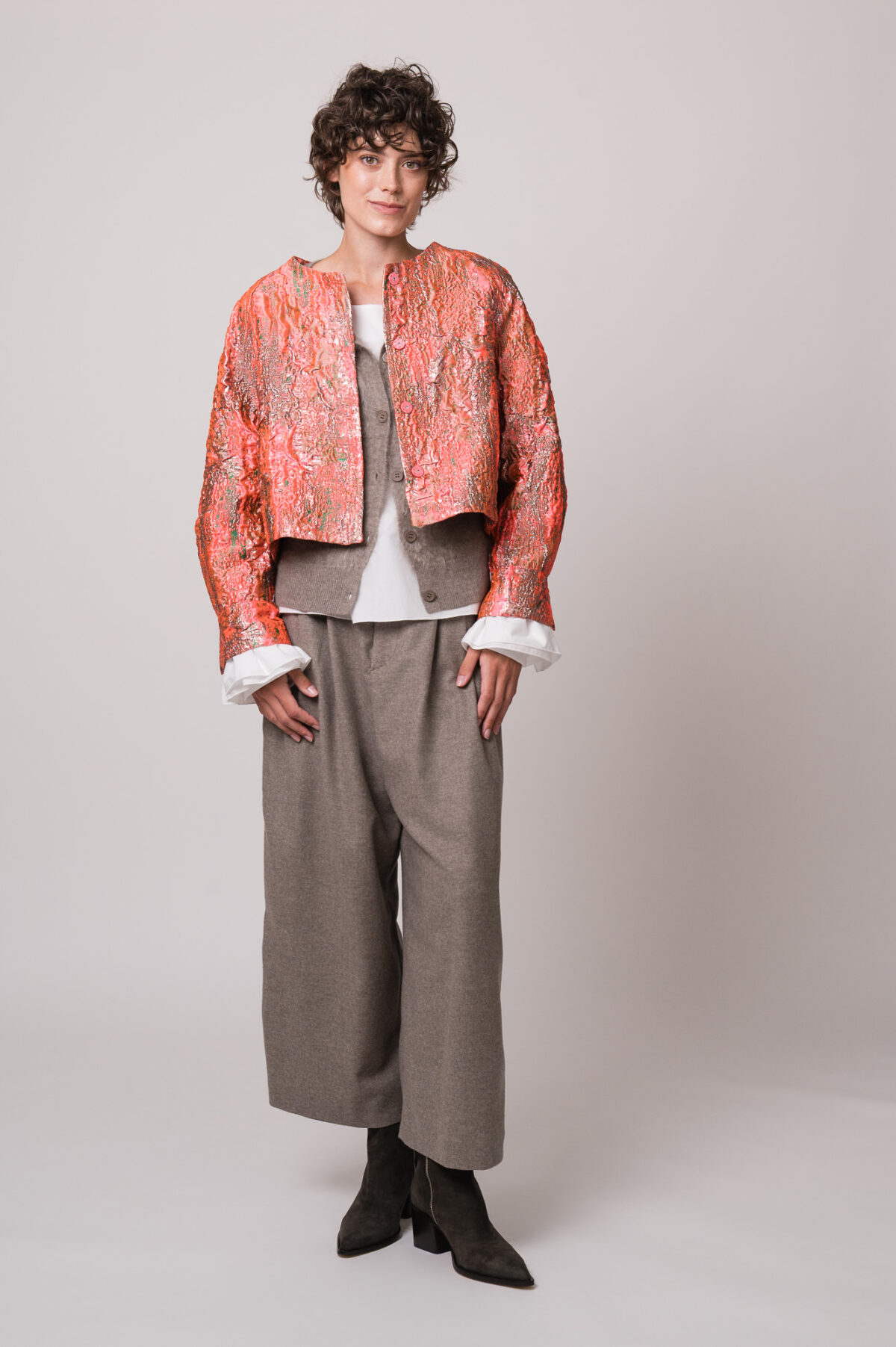 Model trägt eine rosa-goldene Jacke, eine beige Weste, eine weiße Bluse und eine beige Hose der Marke Susanne Bommer.