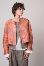 Model trägt eine rosa-goldene Jacke, eine beige Weste, eine weiße Bluse und eine beige Hose der Marke Susanne Bommer.