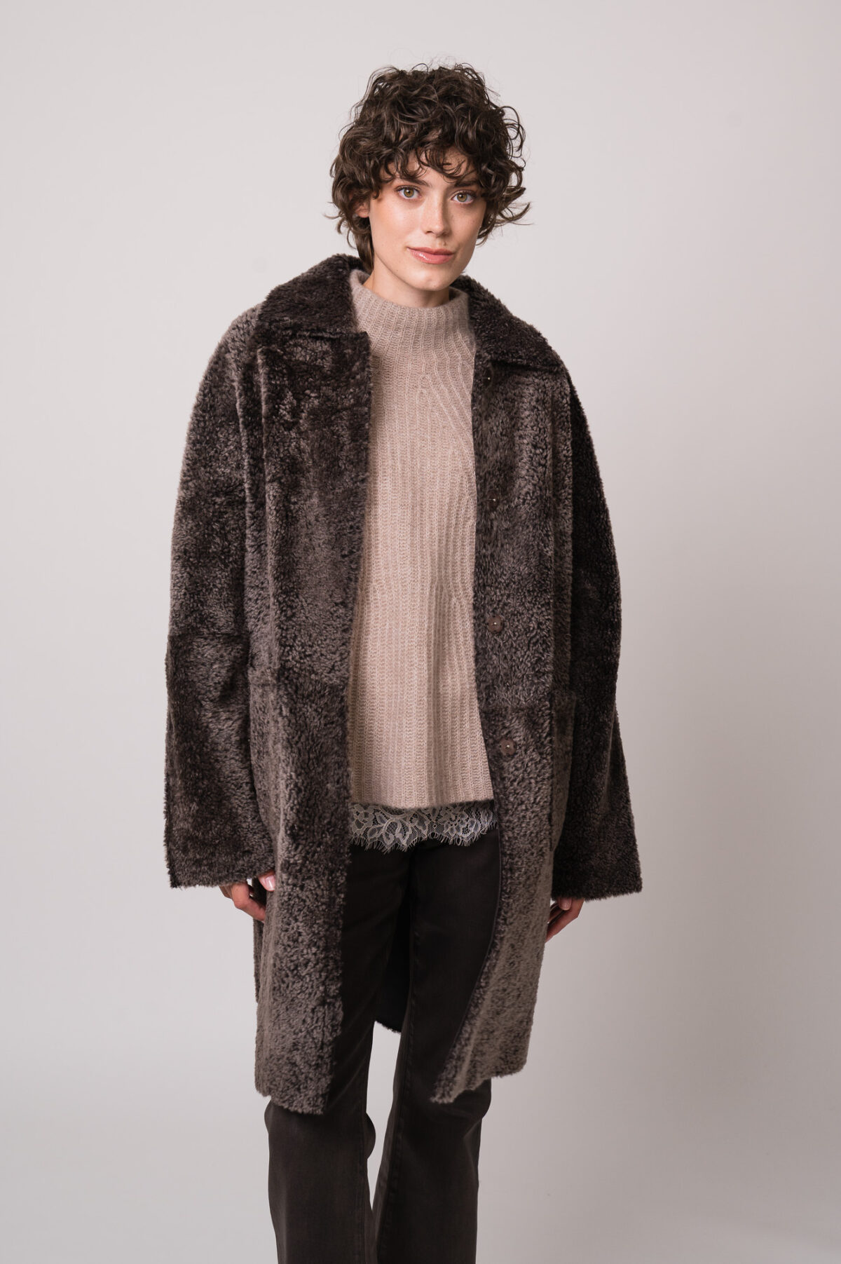 Model trägt einen braunen Lammfellmantel von Hemisphere, eine beiges Top mit Spitze der Marek Allude, einen beigen Pullover der Marke Hemisphere, eine braune Jeans der Marke 7 for all mankind.