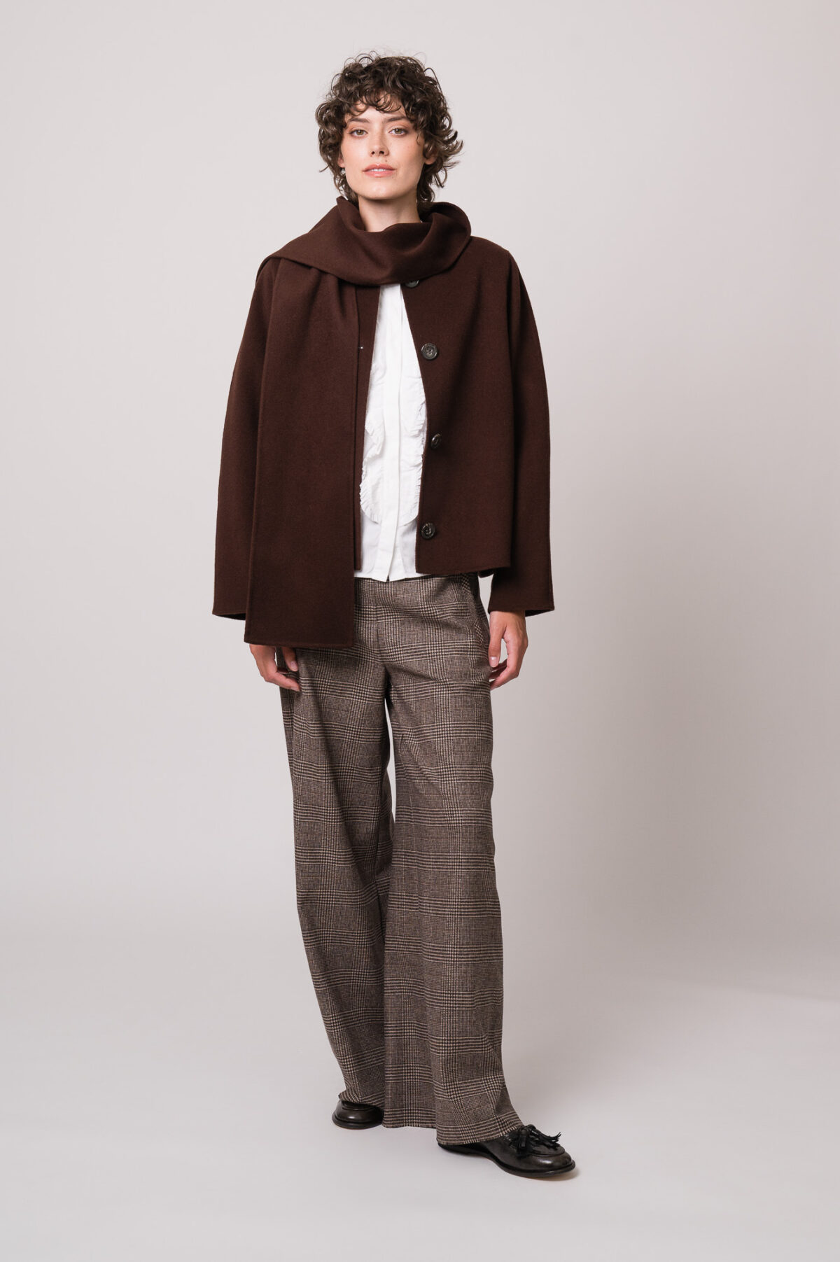 Model trägt eine dunkelbraune Jacke der Marke Luisa Cerano, eine weiße Bluse der Marke Soluzione, eine braun karierte Hose der Marke Cambio.