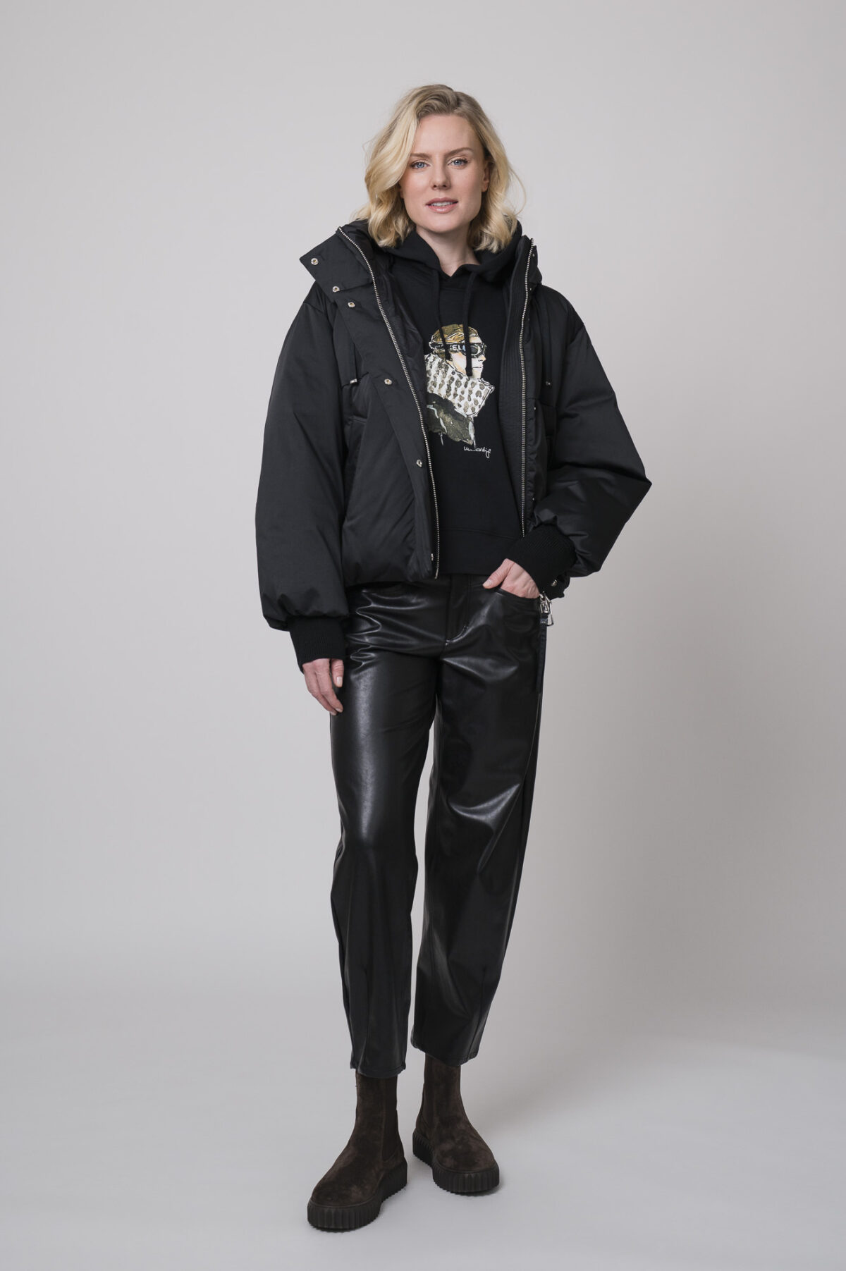 Das Model trägt eine schwarze Daunenjacke der Marke Duno sowie einen Lounge-Pullover von Unartig mit einem Motiv und eine schwarze Hose von Cambio.