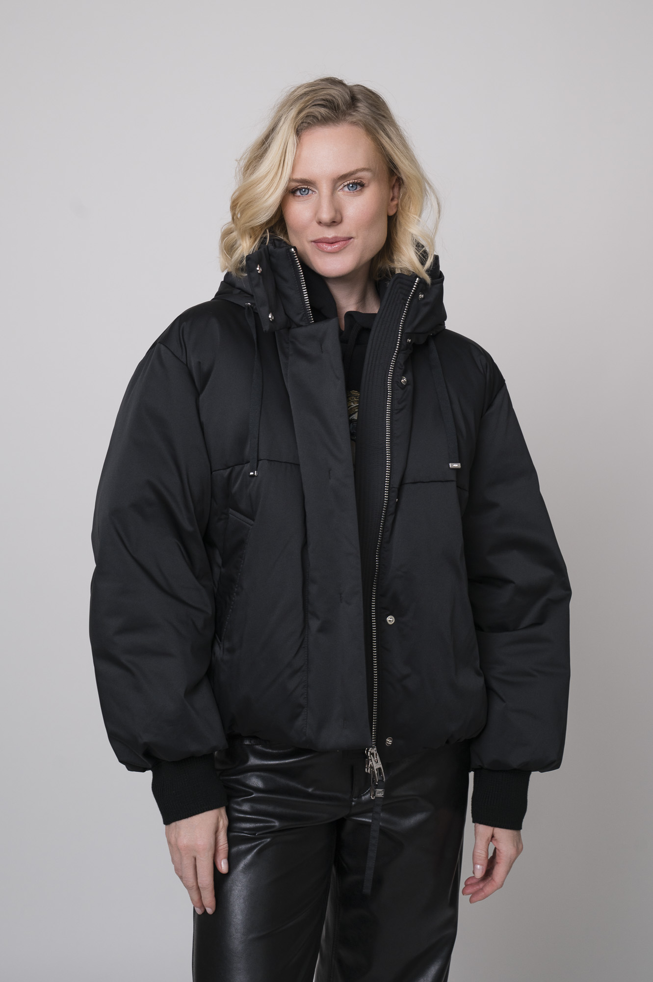 Das Model trägt eine schwarze Daunenjacke der Marke Duno sowie einen Lounge-Pullover von Unartig mit einem Motiv und eine schwarze Hose von Cambio.