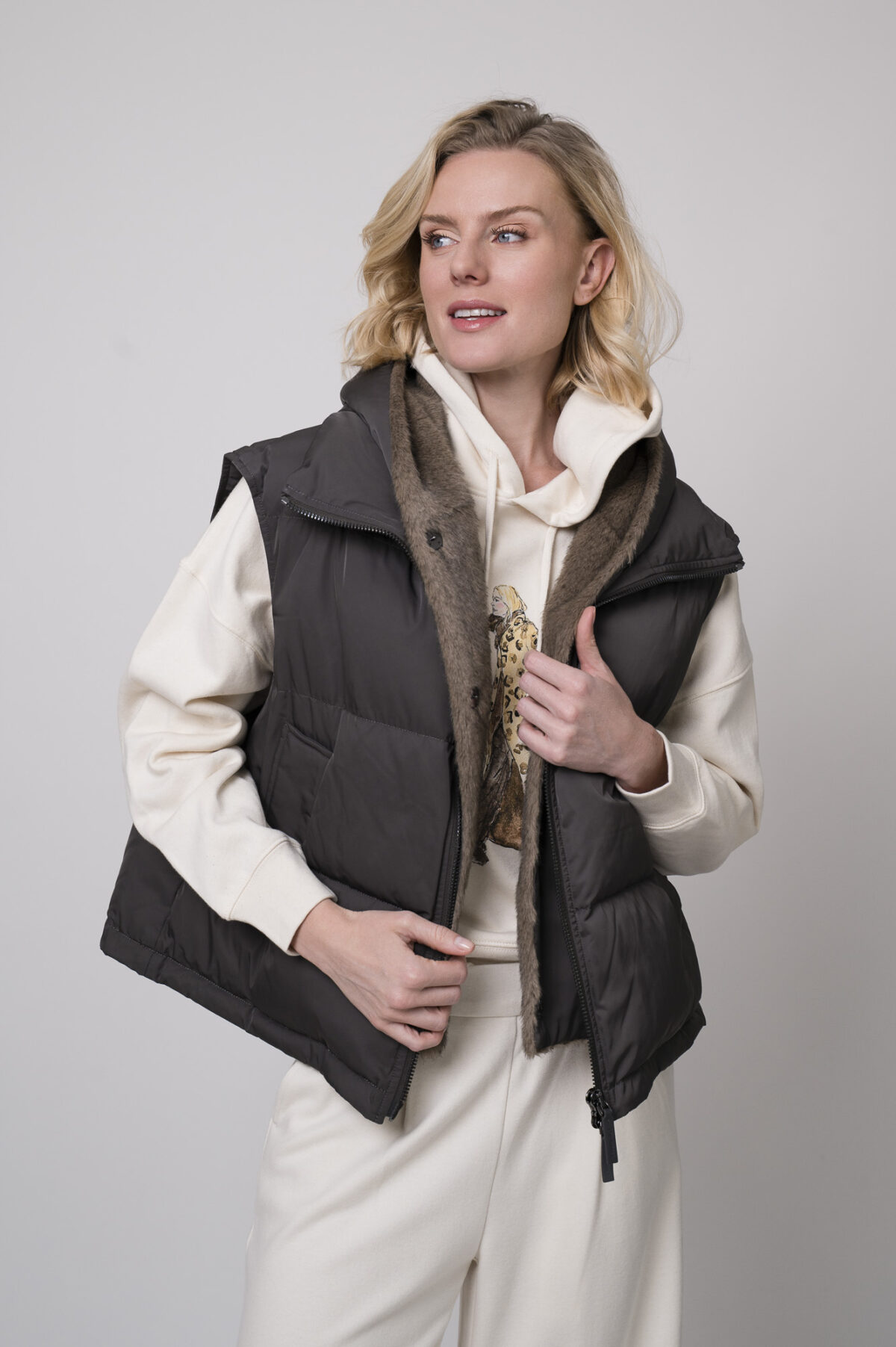 Das Model trägt einen Lounge-Pullover der Marke Unartig mit einem Motiv sowie eine Hose von Cambio in Natur und ein Gilet der Marke Lou Andrea in Anthrazit.