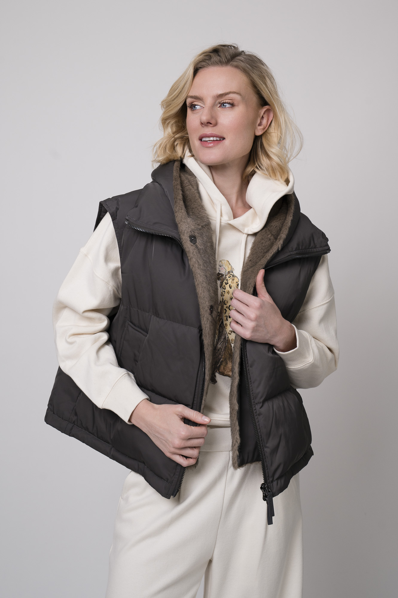 Das Model trägt einen Lounge-Pullover der Marke Unartig mit einem Motiv sowie eine Hose von Cambio in Natur und ein Gilet der Marke Lou Andrea in Anthrazit.