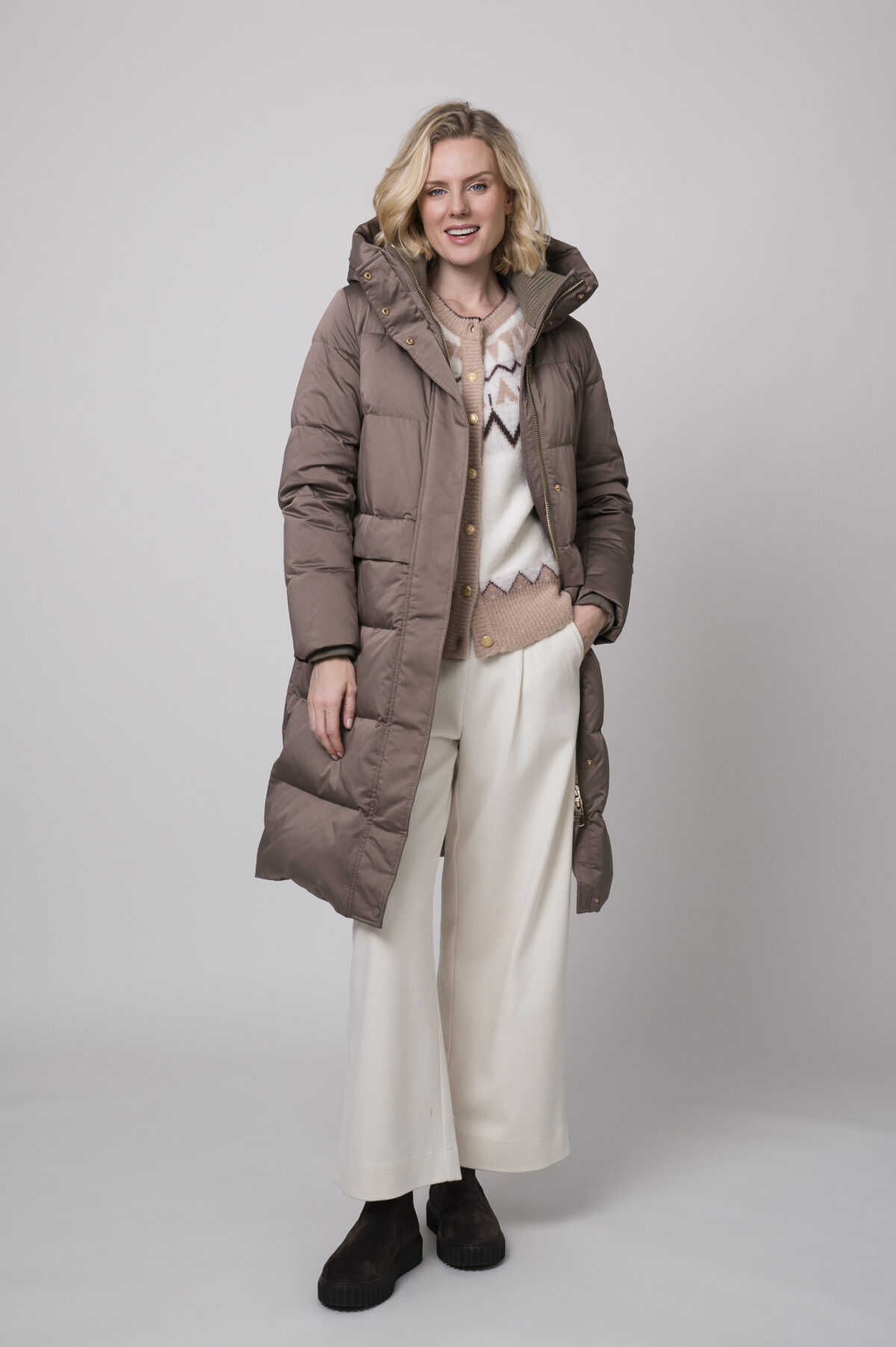Das Model trägt einen braunen Daunenmantel von Duno, eine gemusterte Strickweste von Sosue und eine Hose von Cambio in Natur.