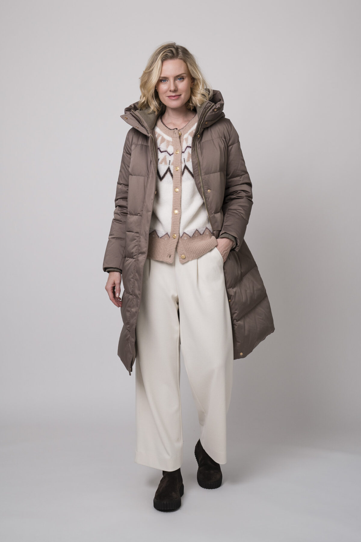 Das Model trägt einen braunen Daunenmantel von Duno, eine gemusterte Strickweste von Sosue und eine Hose von Cambio in Natur.