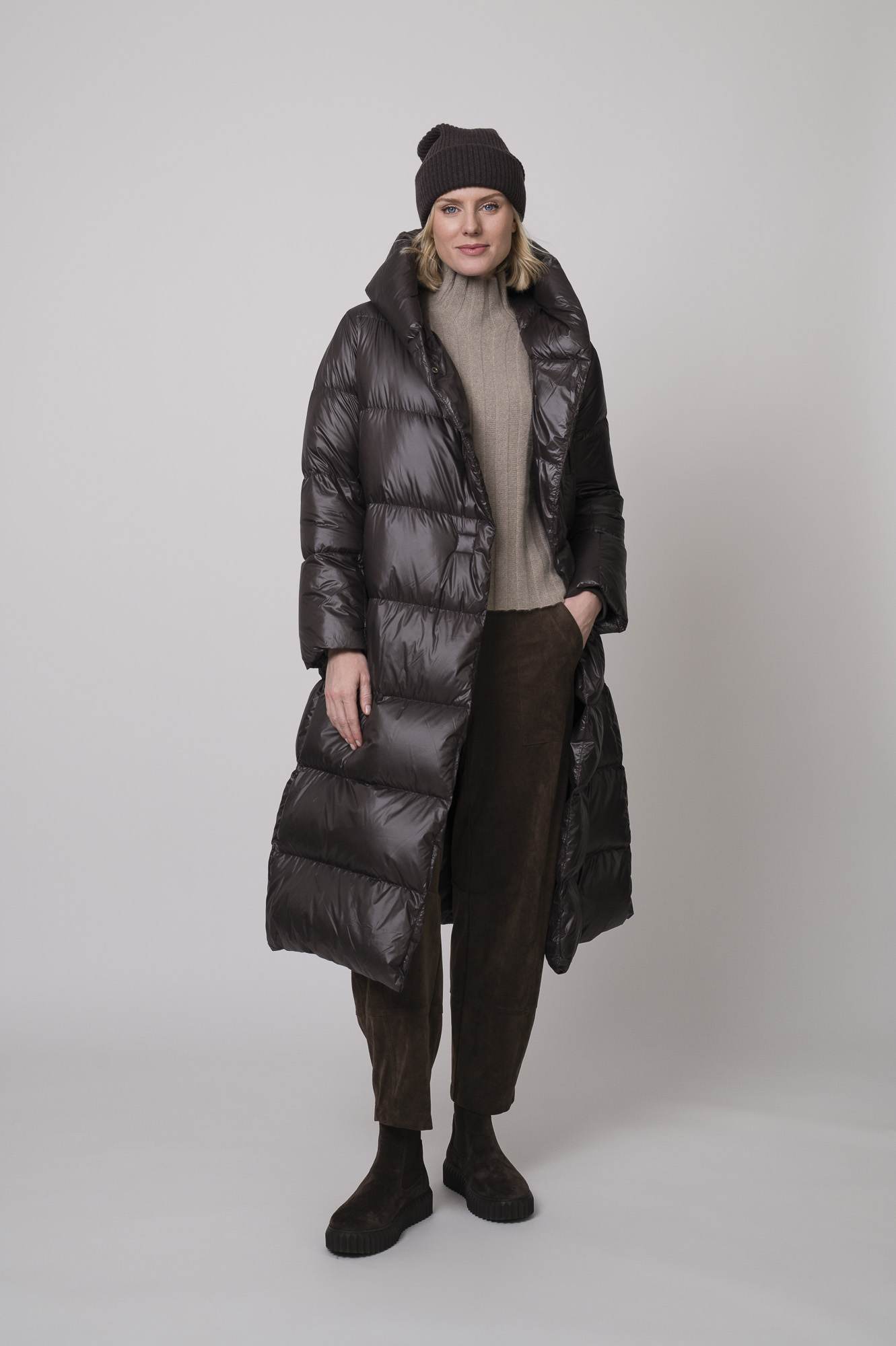 Das Model trägt einen Stehkragenpullover von Hemisphere in Sand, dazu eine braune Hose von Cambio, eine braune Daunenjacke von Helmur und eine Haube von FFC.