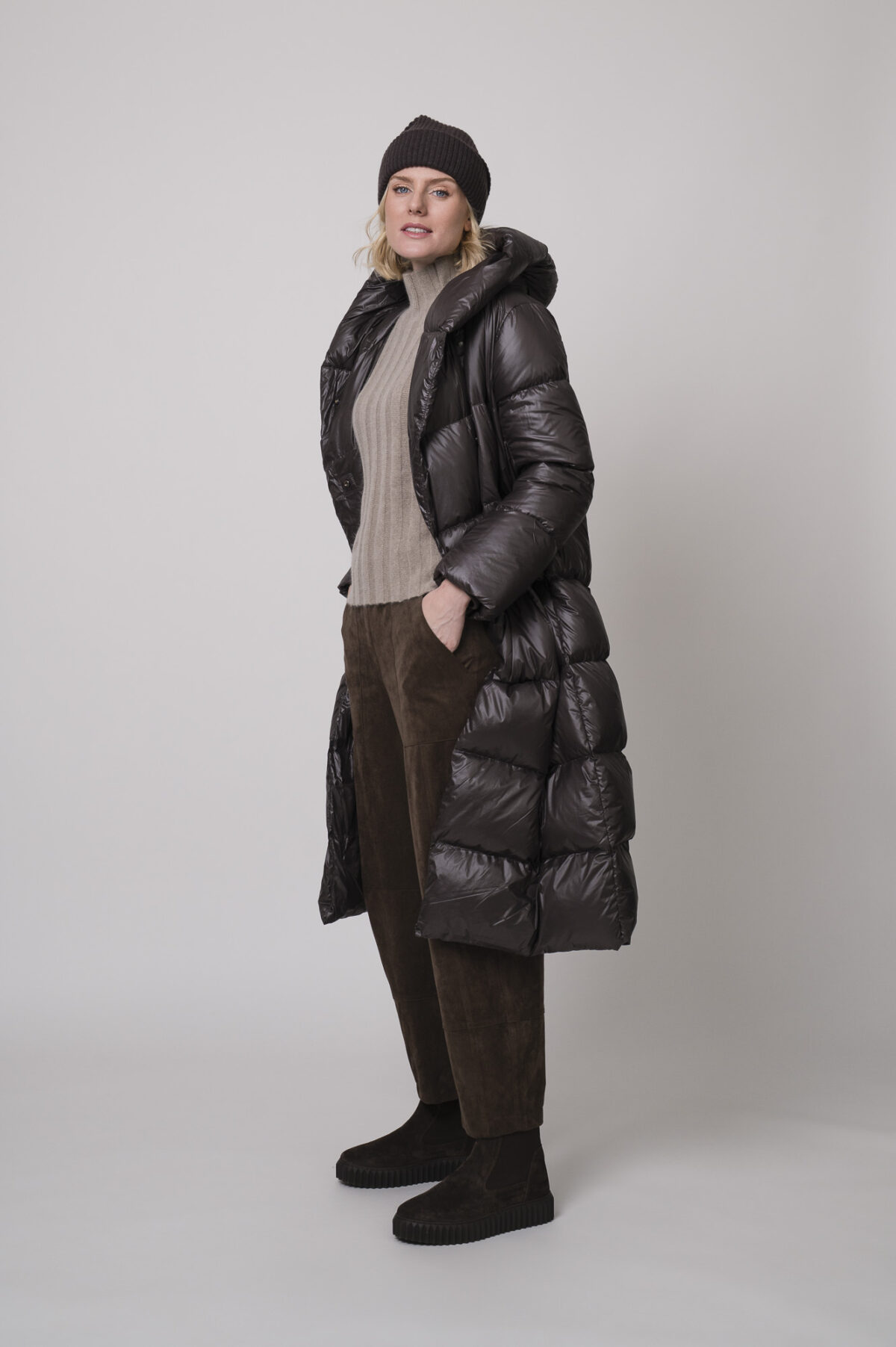 Das Model trägt einen Stehkragenpullover von Hemisphere in Sand, dazu eine braune Hose von Cambio, eine braune Daunenjacke von Helmur und eine Haube von FFC.