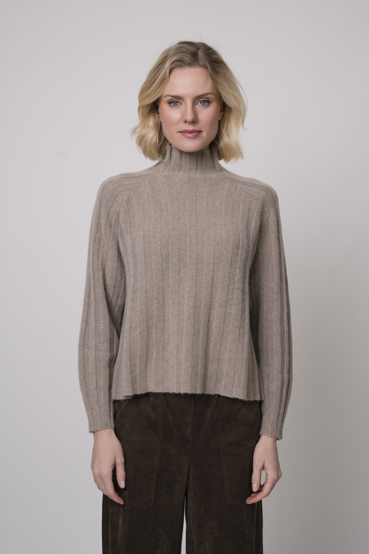 Das Model trägt einen Stehkragenpullover von Hemisphere in Sand, dazu eine Hose in braun von Cambio.
