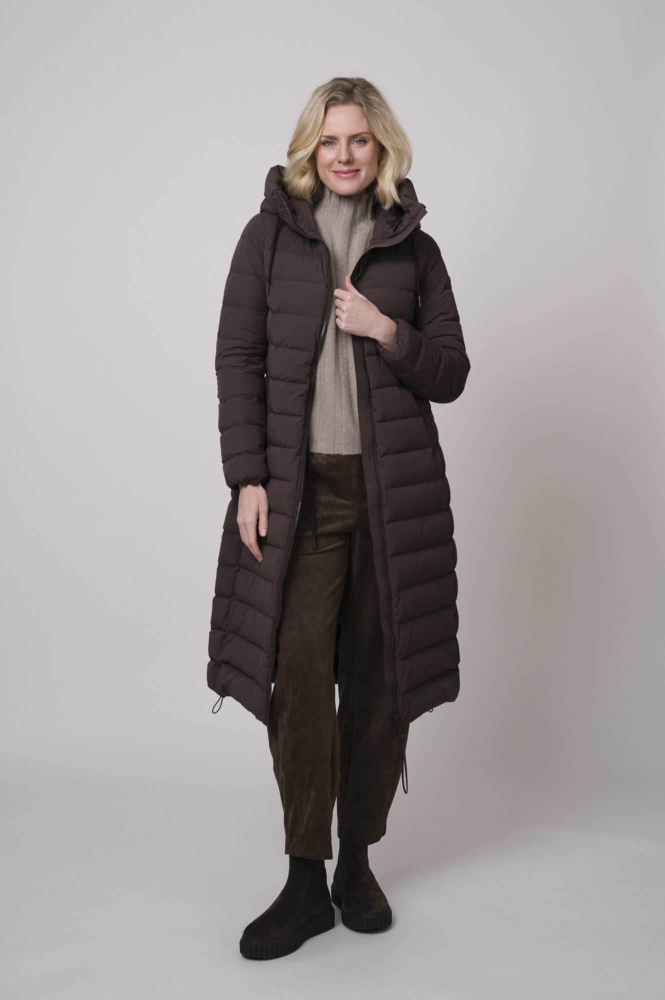 Das Model trägt einen Stehkragenpullover von Hemisphere in Sand, dazu eine Hose in braun von Cambio und einen braunen Mantel der Marke Duno.