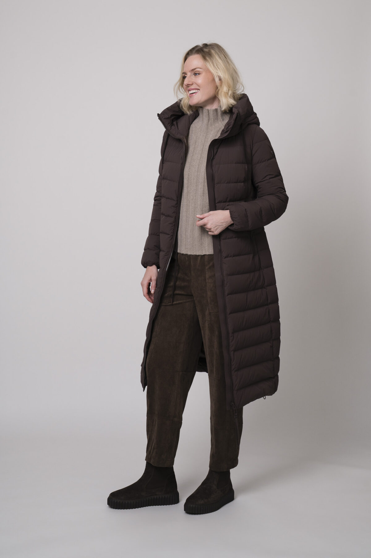Das Model trägt einen Stehkragenpullover von Hemisphere in Sand, dazu eine Hose in braun von Cambio und einen braunen Mantel der Marke Duno.