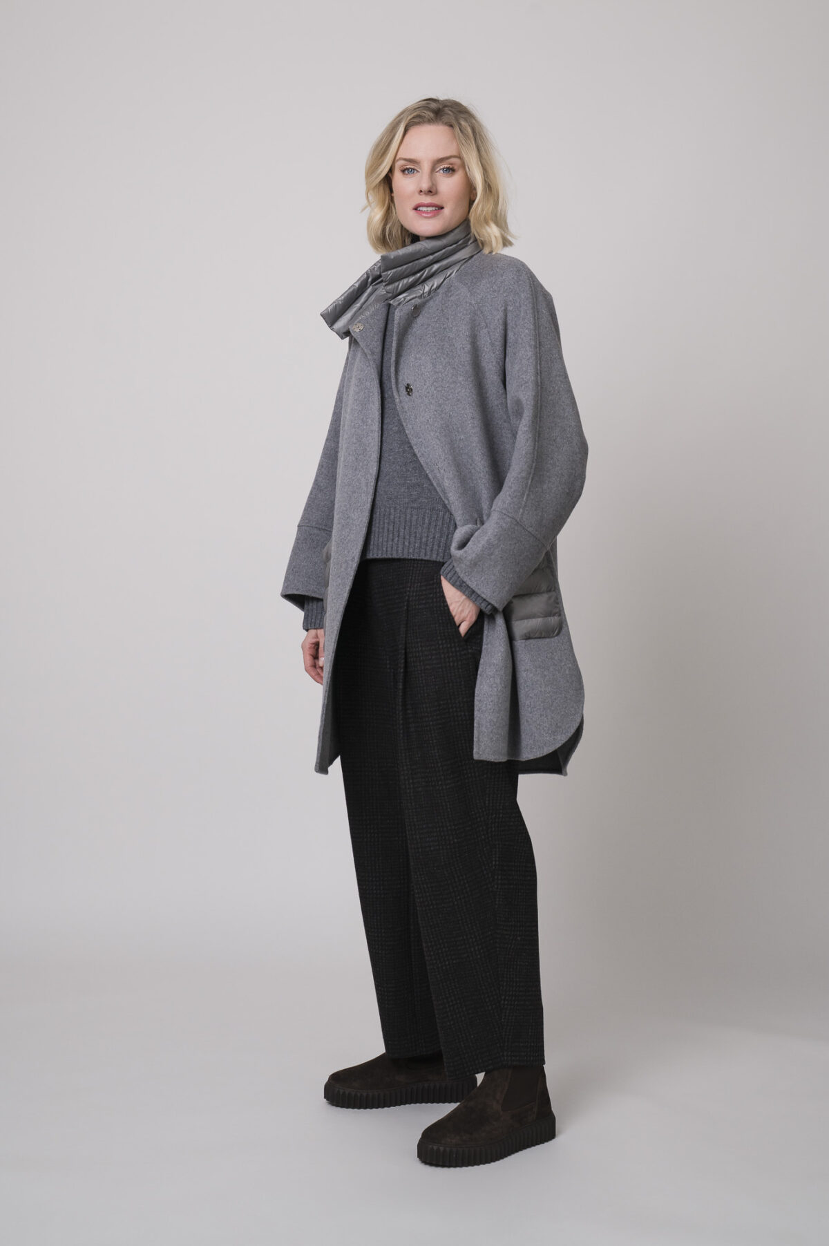 Das Model trägt einen Stehkragenpullover von Luisa Cerano, eine anthrazitfarbene, karierte Hose von Cambio und eine mittelgraue Jacke von Helmur.