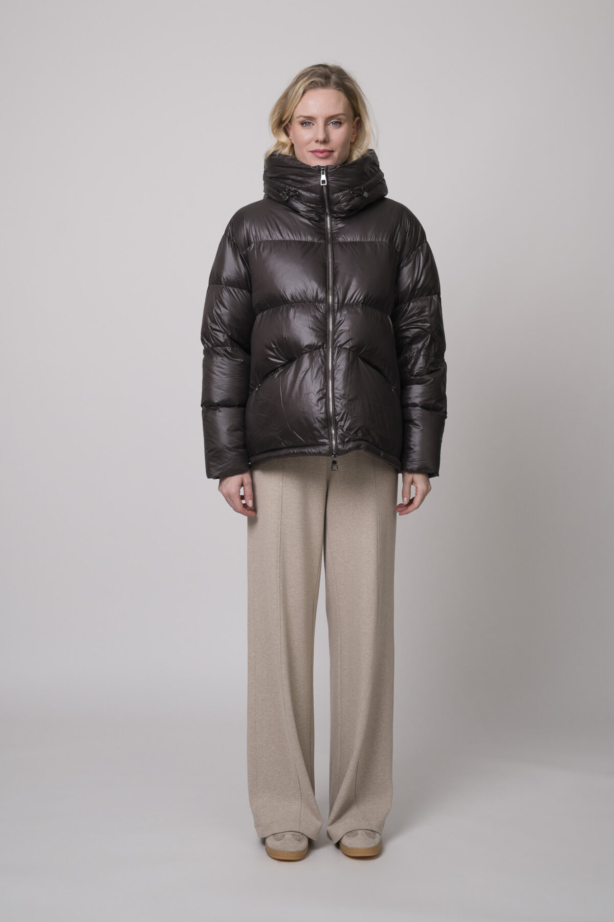 Das Model trägt eine braune Daunenjacke der Marke Helmur, dazu eine Hose der Marke Cambio in Sand und einen braunen Pullover der Marke Sosue.