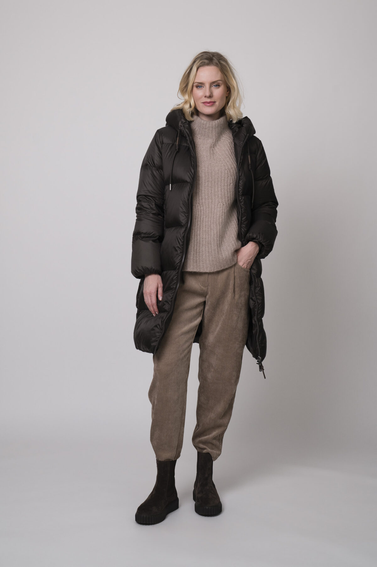 Das Model trägt einen Stehkragenpullover von Hemisphere in Sand, dazu eine Cordhose von Cambio in Camel und einen braunen Daunenmantel von Mabrun.