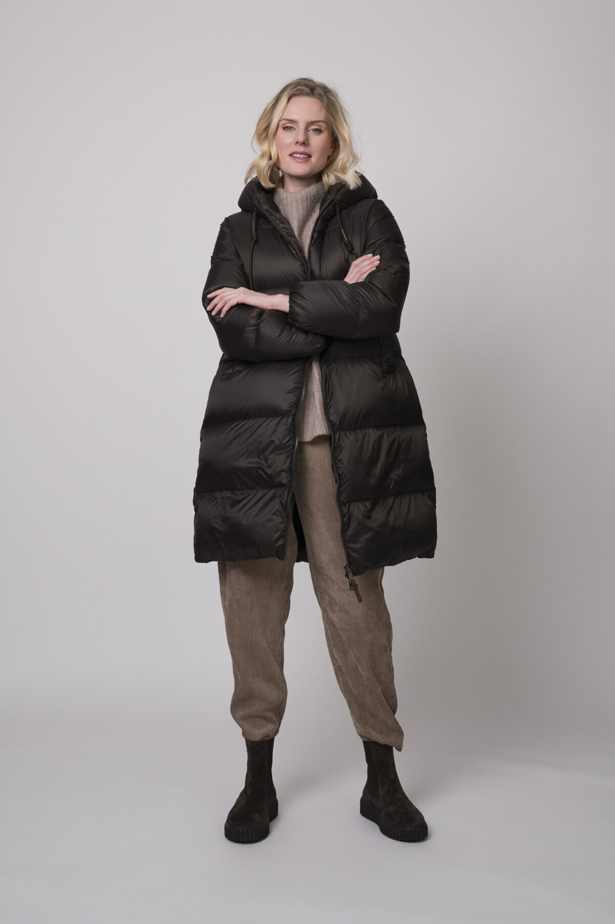 Das Model trägt einen Stehkragenpullover von Hemisphere in Sand, dazu eine Cordhose von Cambio in Camel und einen braunen Daunenmantel von Mabrun.