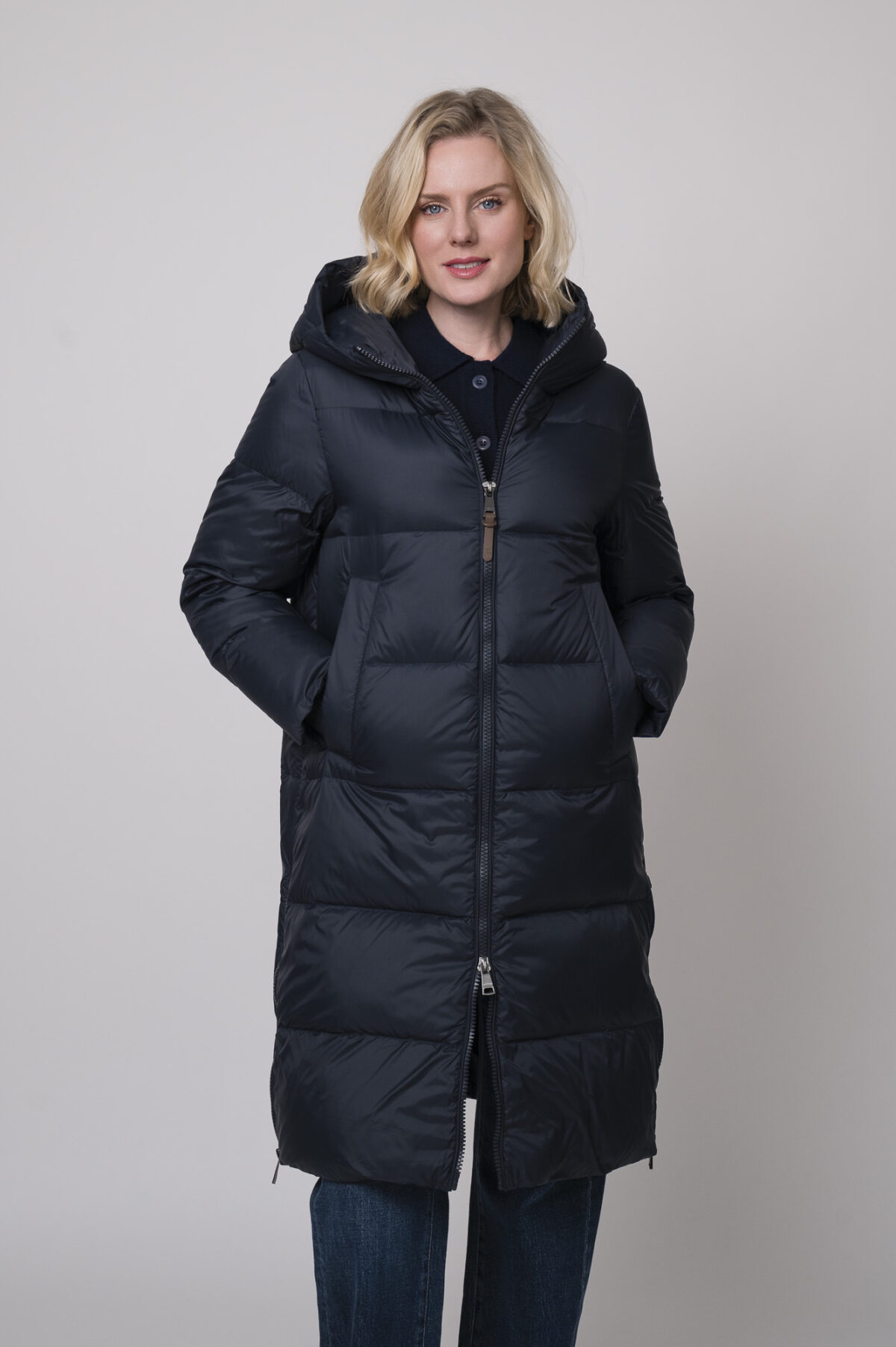 Das Model trägt einen Daunenmantel von Mabrun in Marineblau, eine Jeans von 7 for all mankind und eine Strickweste von True Religion in Marine.