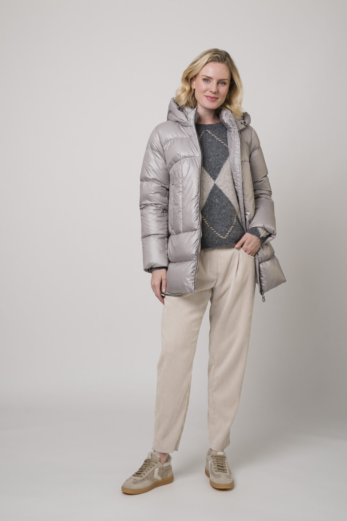Das Model trägt eine Daunenjacke von Helmur in Silber sowie eine Cordhose von Camio in der Farbe Sand und Sneaker von Voile Blanche.