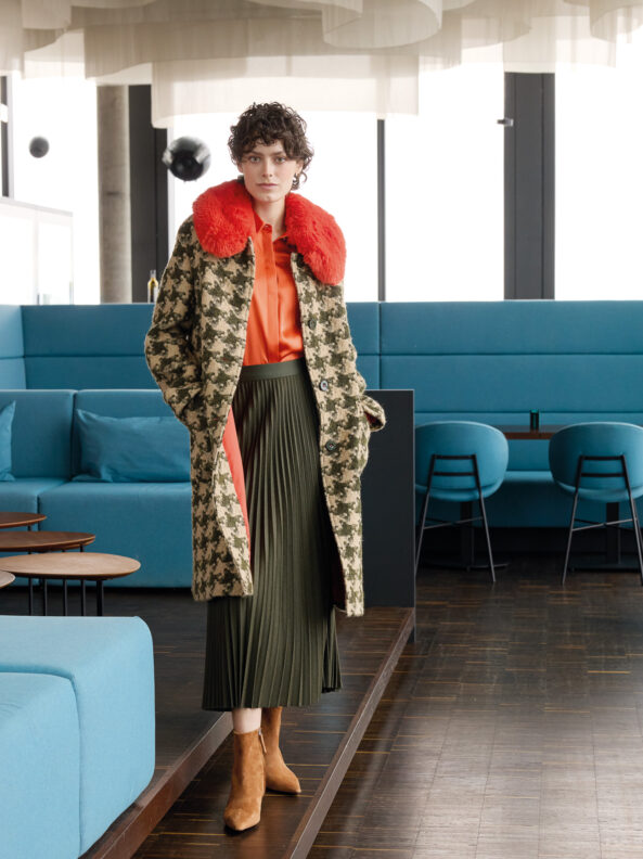 Model trägt ein Outfit von Marc Cain - einen grün gemusterten Mantel mit orangem Kragen, eine orangene Bluse und einen grünen Faltenrock