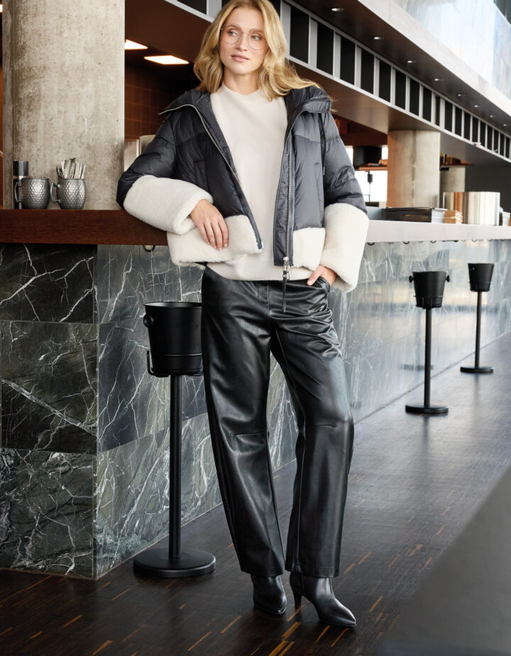 Model trägt eine schwarz-weiße Daunenjacke der Marke Duno, einen weißen Pullover der Marke Herzens und eine schwarze Hose der Marke Cambio