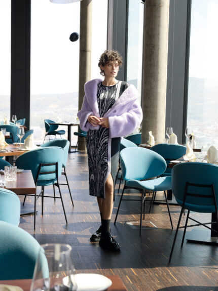 Model trägt ein lila schwarz gemustertes Kleid und eine lila Fake Fur Weste der Marke Marc Cain