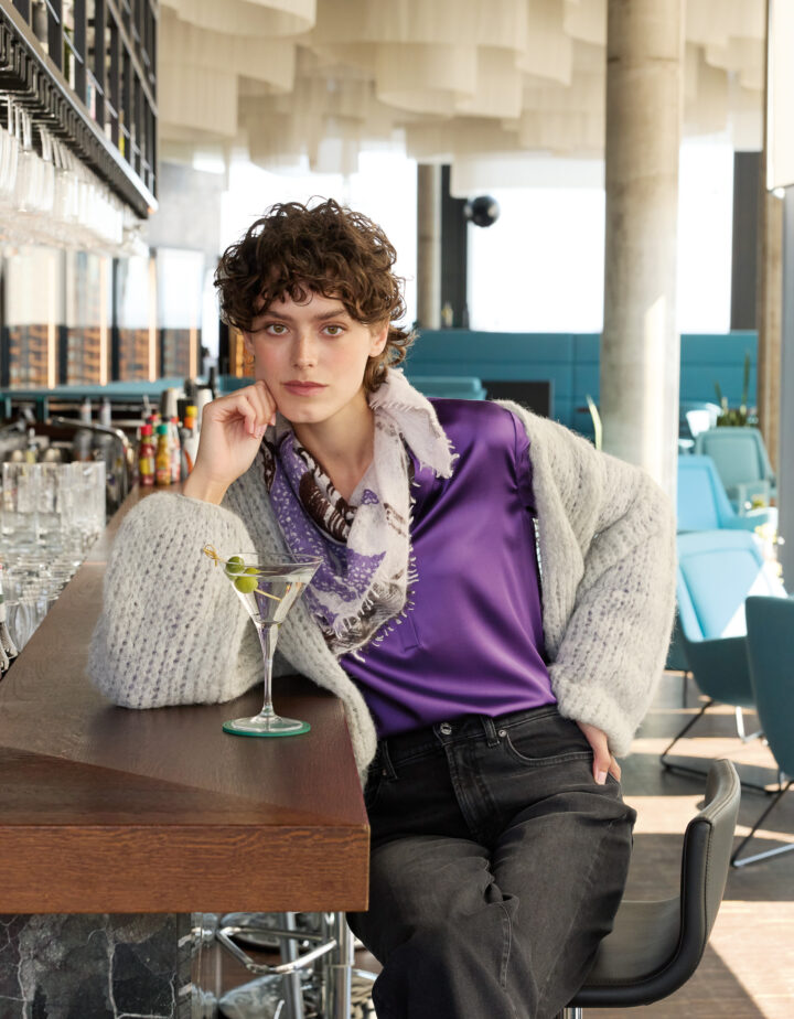 Moel trägt eine graue Strickweste der Marke Michele & Hoven, eine violette Bluse der Marke The Mercer und eine schwarze Jeans der Marke 7 for all Mankind