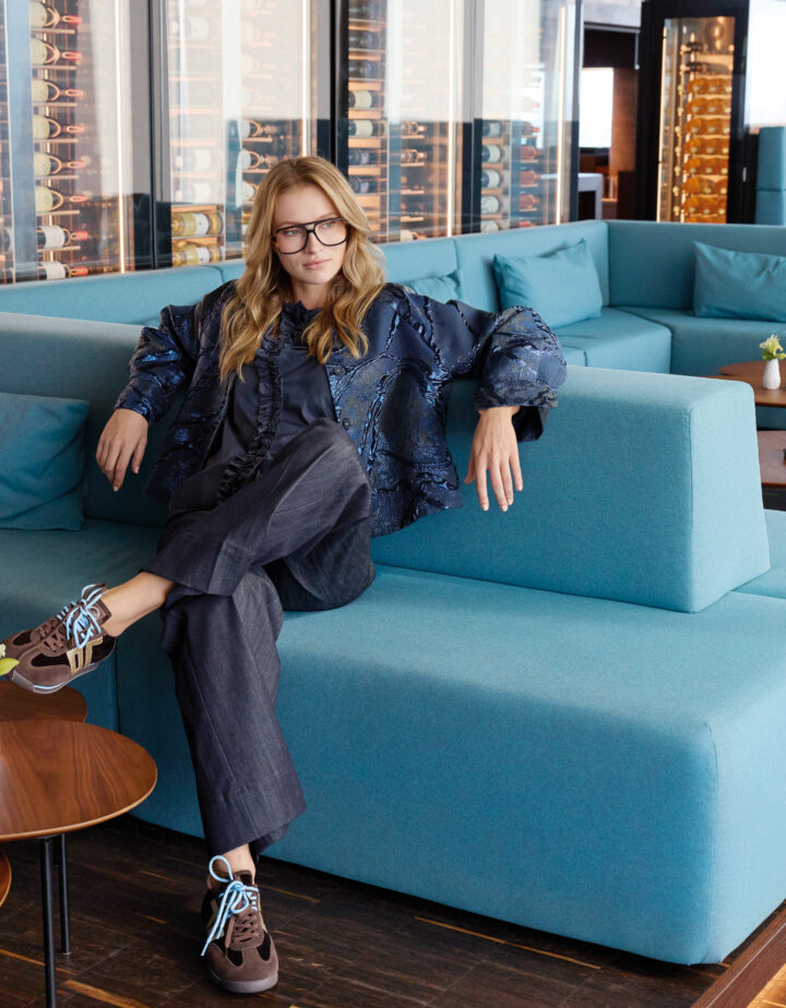 Model trägt eine metallic-blaues Jacke der Marke Susanne Bommer, eine marine-blaue Bluse mit Vollant der Marke Susanne Bommer und blaue Jeans der Marke Mason's